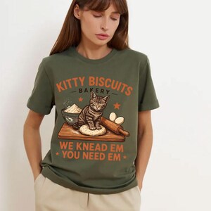 May include: An olive green t-shirt featuring a graphic of a tabby cat on a wooden cutting board with a bowl of flour, eggs, and a rolling pin. The text reads KITTY BISCUITS BAKERY and WE KNEAD EM YOU NEED EM in orange lettering with small stars.