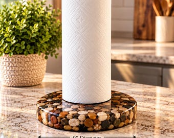 May include: A cylindrical paper towel holder featuring a circular base made of embedded smooth, multi-colored river stones. The base has a 9-inch diameter and supports a standard roll of white paper towels with a diamond-shaped embossed pattern.