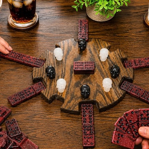 Op de afbeelding: Een rond houten spelbord met ingekeepte randen bevat een centrale dominosteen en zes omliggende schedelvormige speelstukken in zwart en wit. Verschillende donkerrode glitterende dominostenen zijn rond het bord gerangschikt.