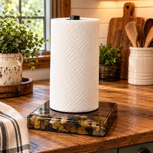 May include: A white paper towel roll stands upright on a square base filled with smooth, multi-colored river stones encased in clear resin. The base features a central black metal rod that secures the paper towel roll in a vertical position.