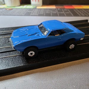 May include: A bright blue toy car with black wheels and silver rims sits on a black track. The car has a classic muscle car design with a sleek, aerodynamic shape. The car is a miniature replica of a vintage vehicle.