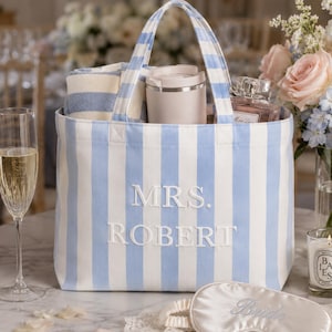 May include: A light blue and white striped canvas tote bag featuring the embroidered text MRS. ROBERT in white. The bag contains a folded striped towel, a beige tumbler, and a glass perfume bottle. A white satin sleep mask with the word Bride is in front.