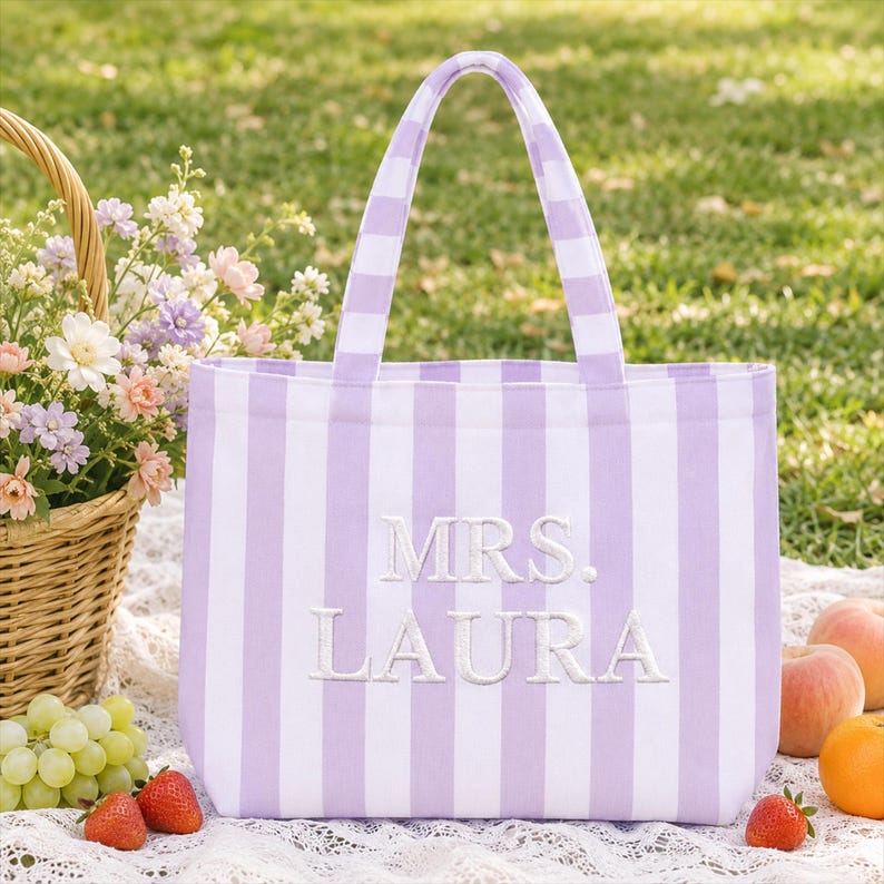 May include: A tote bag featuring vertical lavender and white stripes with the text MRS. LAURA embroidered in white thread on the front. The bag has two matching striped handles and a rectangular structure made of fabric.