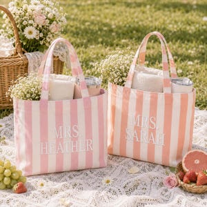 May include: Two rectangular canvas tote bags with vertical stripes. The left bag is pink and white with the embroidered text "MRS. HEATHER". The right bag is peach and white with the embroidered text "MRS. SARAH". Both bags contain white towels and tumblers.