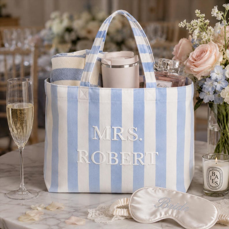 May include: A light blue and white striped canvas tote bag featuring the embroidered text MRS. ROBERT in white. The bag contains a folded striped towel, a beige tumbler, and a glass perfume bottle. A white satin sleep mask with the word Bride is in front.