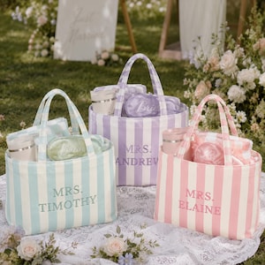 May include: Three striped canvas tote bags in light blue, lavender, and pink. Each bag features embroidered text reading MRS. TIMOTHY, MRS. ANDREW, or MRS. ELAINE. Inside each bag are matching sleep masks and tumblers with lids, arranged in a set.