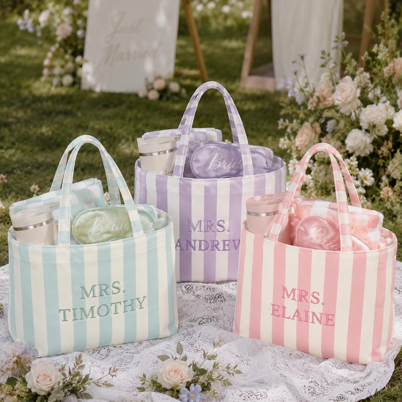 May include: Three striped canvas tote bags in light blue, lavender, and pink. Each bag features embroidered text reading MRS. TIMOTHY, MRS. ANDREW, or MRS. ELAINE. Inside each bag are matching sleep masks and tumblers with lids, arranged in a set.