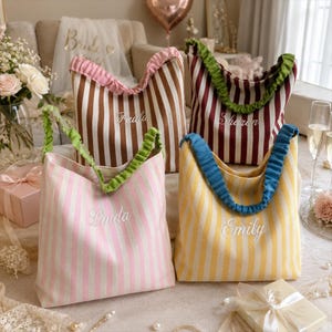 Può includere: Quattro borse in tessuto a righe verticali marroni, bordeaux, rosa e gialle. Ogni borsa presenta un manico arricciato e nomi ricamati in bianco: Paula, Sharon, Linda ed Emily. Le borse hanno un aspetto morbido e strutturato.