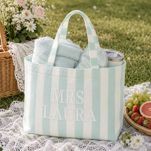May include: A light mint green and white striped canvas tote bag featuring the embroidered text MRS. LAURA on the front. The bag contains a folded light green towel, a matching eye mask, and a silver-toned tumbler with a clear lid.