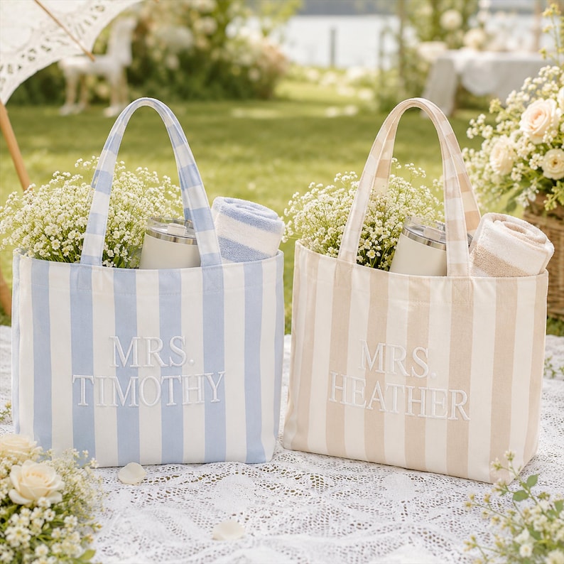May include: Two canvas tote bags with vertical stripes, one in light blue and white, the other in beige and white. Each bag features embroidered text, with the blue bag reading MRS. TIMOTHY and the beige bag reading MRS. HEATHER, filled with flowers.