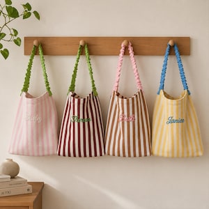 Pode incluir: Quatro sacolas listradas penduradas em um cabideiro de madeira. As bolsas apresentam nomes bordados em cursivo: Shirley em uma bolsa rosa, Theresa em uma bordô, Beth em uma marrom e Janice em uma amarela. Cada bolsa tem uma alça franzida.