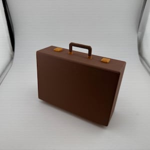 May include: A rectangular brown briefcase with a textured surface, featuring a dark brown top handle and two small yellow latches on the upper edge. The case has a structured, boxy design with clean lines and a solid, uniform color throughout the exterior.
