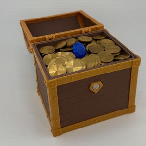 Könnte beinhalten: Eine braune Schatzkiste mit einem Klappdeckel, gefüllt mit Goldmünzen und einem blauen Edelstein. Die Truhe hat eine rechteckige Form mit goldfarbenen Verzierungen und Nieten. Das Innere ist mit zahlreichen runden Goldmünzen gefüllt.