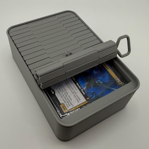 Puede incluir: Una caja de almacenamiento de tarjetas de plástico gris con una tapa con bisagras y un asa. La tapa está parcialmente abierta, revelando una pila de cartas coleccionables. Las tarjetas tienen un diseño gráfico azul y negro y el texto "Horror Pirate".