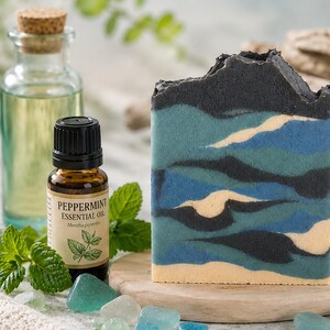 May include: A rectangular bar of soap featuring a layered, wavy pattern in shades of black, teal, blue, and cream. Beside it sits a small amber glass bottle labeled Peppermint Essential Oil with a black cap, fresh mint leaves, and sea glass.