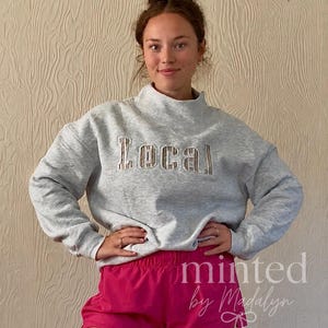 May include: A light gray mock neck sweatshirt featuring the word Local in a tan and white striped fabric appliqué across the chest, paired with bright pink athletic shorts. The sweatshirt has long sleeves and a relaxed fit.