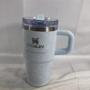 May include: A light blue Stanley tumbler with a white floral pattern, featuring the Stanley logo and the text LOVESHACKFANCY printed on the front. The tumbler has a clear plastic lid with a light blue tab and a sturdy light blue side handle.