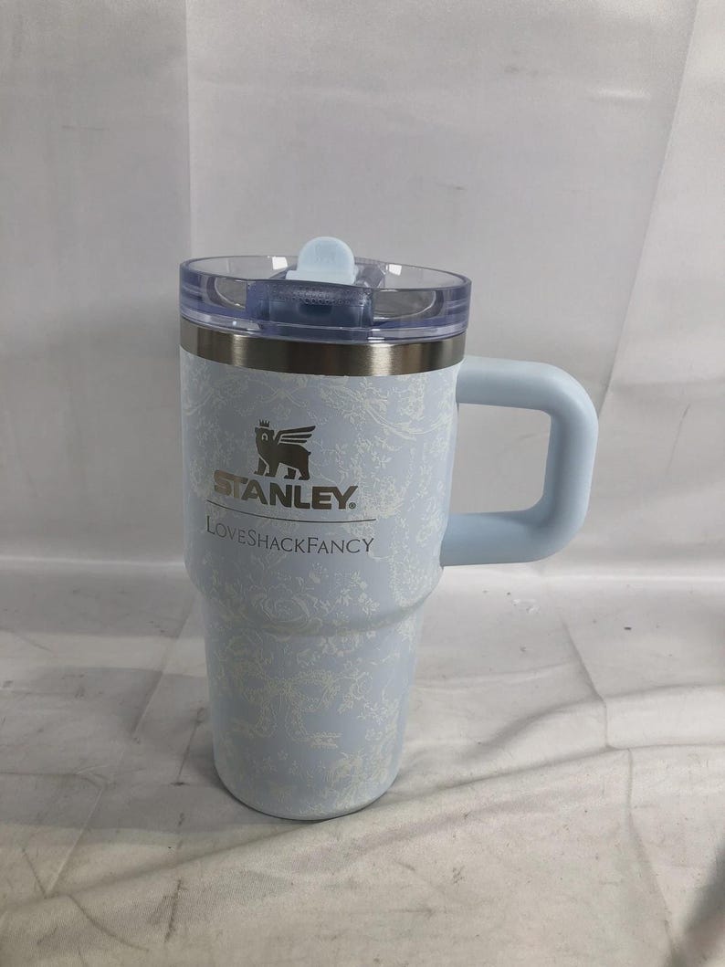 May include: A light blue Stanley tumbler with a white floral pattern, featuring the Stanley logo and the text LOVESHACKFANCY printed on the front. The tumbler has a clear plastic lid with a light blue tab and a sturdy light blue side handle.