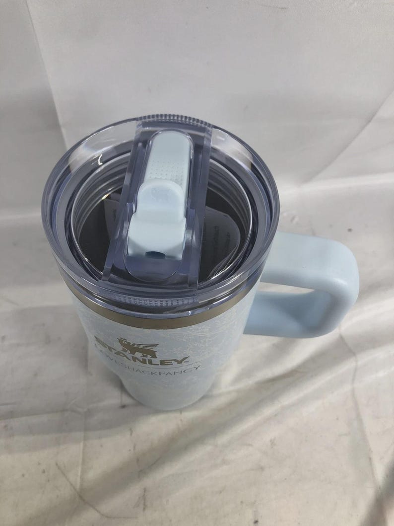 Puede incluir: Un vaso Stanley LoveShackFancy de color azul claro con tapa de pl&aacute;stico transparente y asa azul claro. El vaso presenta una superficie texturizada y el logotipo de Stanley LoveShackFancy impreso en oscuro en el lateral. La tapa tiene un mecanismo deslizante.