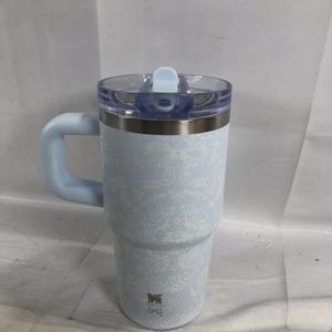 Puede incluir: Un vaso t&eacute;rmico de color azul claro con un patr&oacute;n de damasco blanco, que incluye un asa a juego, una tapa de pl&aacute;stico transparente con cierre deslizante azul claro y un peque&ntilde;o logotipo cerca de la base de forma cil&iacute;ndrica.