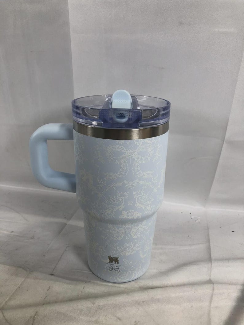 Puede incluir: Un vaso t&eacute;rmico de color azul claro con un patr&oacute;n de damasco blanco, que incluye un asa a juego, una tapa de pl&aacute;stico transparente con cierre deslizante azul claro y un peque&ntilde;o logotipo cerca de la base de forma cil&iacute;ndrica.