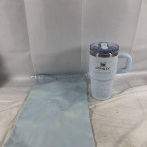 May include: A light blue Stanley tumbler with a handle and a clear lid, featuring a subtle white floral pattern and the text Stanley LoveShackFancy printed on the front. Beside it is a matching light blue drawstring fabric storage bag.