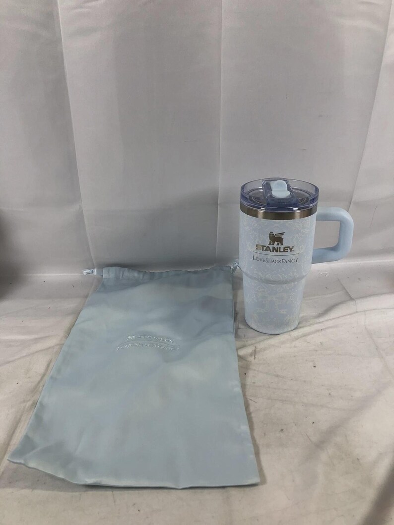 May include: A light blue Stanley tumbler with a handle and a clear lid, featuring a subtle white floral pattern and the text Stanley LoveShackFancy printed on the front. Beside it is a matching light blue drawstring fabric storage bag.