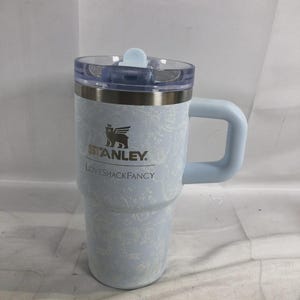 Puede incluir: Un vaso Stanley de color azul claro con un estampado floral blanco, que presenta el logotipo de Stanley y el texto LOVESHACKFANCY impreso en la parte frontal. El vaso tiene una tapa de pl&aacute;stico transparente con una pesta&ntilde;a azul y asa lateral.