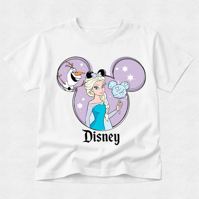 May include: A white short-sleeve t-shirt featuring a graphic of Elsa and Olaf inside a purple Mickey Mouse ear silhouette. Elsa wears black mouse ears and holds a light blue cotton candy treat. The word Disney is printed in black below the graphic.