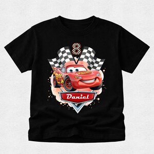 Peut inclure: T-shirt noir avec une voiture de course rouge de dessin anim&eacute;, le num&eacute;ro 95, et un drapeau &agrave; damier. Le design comprend le nom "Daniel" sur une banni&egrave;re et le chiffre 8. La voiture a un visage souriant.