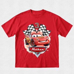 Peut inclure: T-shirt rouge avec un dessin de Flash McQueen du film Cars. Le motif comprend un drapeau &agrave; damier, le chiffre 3 et le nom "Nathan". La voiture est rouge avec des d&eacute;tails jaunes.