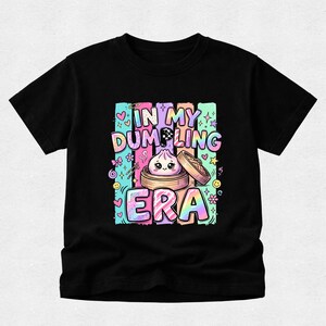 May include: A black short-sleeved t-shirt featuring a colourful graphic print with the text "IN MY DUMPLING ERA". The design includes a cute cartoon dumpling in a bamboo steamer, surrounded by pastel-coloured stripes, hearts, stars, and flowers.