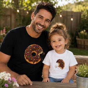 May include: A black t-shirt featuring a large chocolate-frosted doughnut with colourful sprinkles and a bite taken out of it, paired with a white t-shirt featuring a smaller matching chocolate-frosted doughnut graphic with colourful sprinkles.