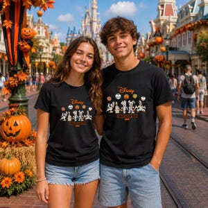 以下が含まれることがあります： Disney Halloween 2026というテキストがプリントされた黒いTシャツ。コスチュームを着た漫画のキャラクターのイラストと、カボチャやキャラクターの顔が描かれた小さな円形のアイコンがデザインされています。