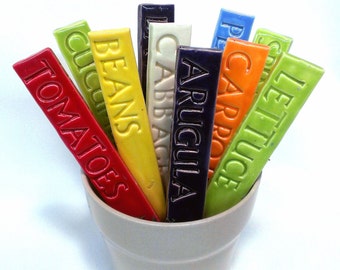 May include: A selection of vibrant ceramic plant markers in a beige pot. Each marker is rectangular and displays embossed text denoting different vegetables, such as "Tomatoes," "Beans," and "Lettuce." The markers are in various colours, including red, yellow, green, and blue.