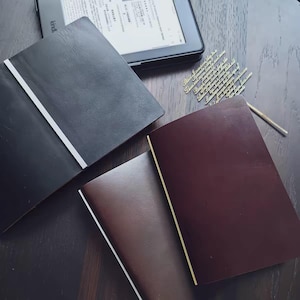 May include: Three leather-bound journals in black, dark brown, and deep burgundy colors, each featuring a thin vertical elastic band. A black e-reader is positioned at the top, and a gold-toned metal bookmark with intricate script text is included.
