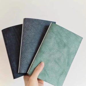 May include: Three rectangular notebooks with textured covers in shades of dark navy, charcoal gray, and sage green. Each notebook features a clean, straight spine and a distinct pebbled surface texture, arranged in a fan shape for clear visibility.