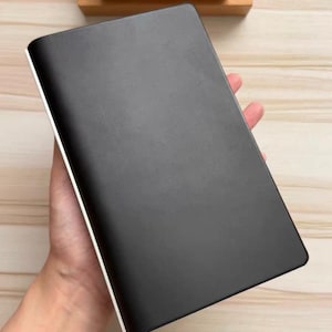 May include: A rectangular black notebook with a smooth matte cover and a thin white edge along the side. The notebook is held in a vertical position, displaying a solid dark surface with rounded corners and a clean, minimalist design aesthetic.