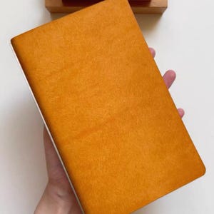 May include: A rectangular notebook with a textured, mustard-yellow cover. Positioned above the notebook is a wooden stand holding three individual wooden blocks, each engraved with a single digit: the numbers 1, 1, and 2 in sequence.