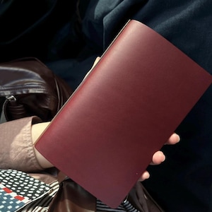 May include: A rectangular, deep burgundy leather notebook or journal cover with a smooth, matte finish. The object is held in a vertical orientation, showcasing a clean, minimalist front surface with no visible branding, stitching, or additional hardware.