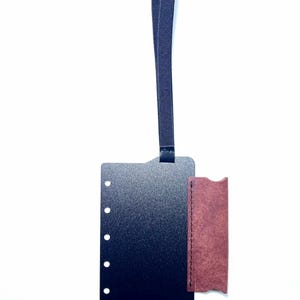 May include: A rectangular black luggage tag with a textured finish and five circular holes along the left edge. A dark blue elastic strap is attached to the top. A small, rectangular, reddish-brown leather piece is stitched to the right side.