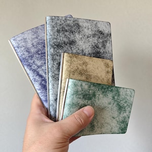 May include: A set of four rectangular wallets or card holders featuring a textured, mottled finish in shades of blue, gray, gold, and green. Each item has a smooth, metallic-toned spine and a distinct, fibrous, or marbled surface pattern.