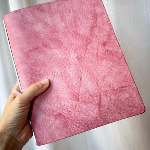 May include: A rectangular notebook with a textured, mottled pink cover featuring a marbled appearance. The cover has a smooth, matte finish with subtle variations in color intensity across the surface, creating a distinct, organic pattern.