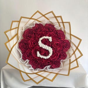 Peut inclure: Un bouquet de roses artificielles rouge foncé disposées en grappe circulaire. Une lettre S blanche, composée de petites fleurs blanches, est centrée sur les roses. Le bouquet est enveloppé dans des couches de papier blanc translucide à bords dorés.