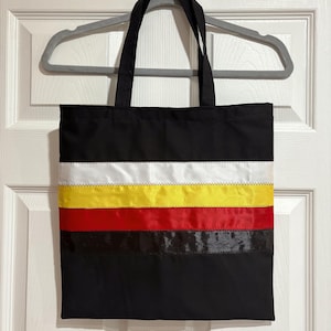 May include: Black tote bag with a horizontal ribbon design in white, yellow, red, and black. The bag has two black handles and is hanging on a gray hanger. The bag is made of a sturdy fabric.