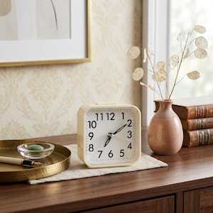 May include: A square analog alarm clock with a cream-colored frame and a white face featuring black Arabic numerals. The clock has two thin, dark-colored hands and a second hand, all centered on the dial, designed for tabletop placement.