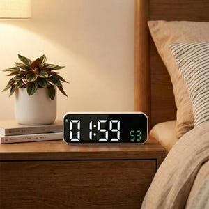 May include: A rectangular digital alarm clock with a black face and white LED display showing the time 01:59 and green seconds 53. The clock has a white frame and is positioned on a wooden surface next to a potted plant and two books.