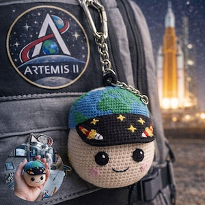 May include: A crocheted keychain featuring a smiling character with a blue and green globe-shaped hat and a black visor with yellow stars and rockets. The keychain is attached to a backpack with an Artemis II patch.