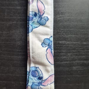 Könnte beinhalten: Ein Schlüsselanhänger aus Stoff mit weißem Hintergrund und einem sich wiederholenden Muster der blauen außerirdischen Figur Stitch. Das Band ist unten mit einer rechteckigen silberfarbenen Metallklemme und einem passenden Metall-D-Ring befestigt.