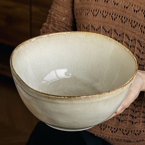 Peut inclure: Un grand bol en céramique rond avec un émail intérieur et extérieur blanc cassé lisse. Le bord présente une finition en émail réactif brun plus foncé distincte qui crée une bordure subtile et texturée autour du haut du récipient.